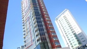 Een hoog, modern gebouw met een gevel in rood en wit, meerdere balkons en een helderblauwe lucht op de achtergrond.