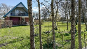 Uma casa moderna com telhado inclinado e uma varanda grande, cercada por um gramado bem cuidado e árvores.