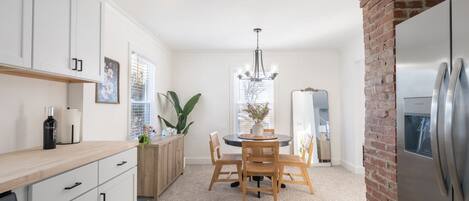Una cocina moderna con muebles blancos, una mesa de comedor con sillas, una araña y una pared de ladrillo decorativa.