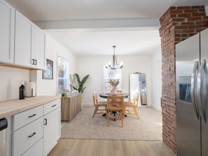 A modern kitchen with white cabinetry, a dining table with chairs, a chandelier, and a brick accent wall.
