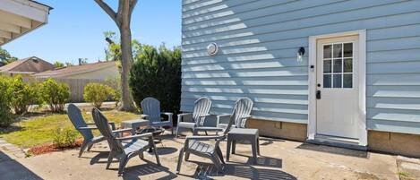 A patio with Adirondack chairs and a small table, a white door, and a tree in the background.