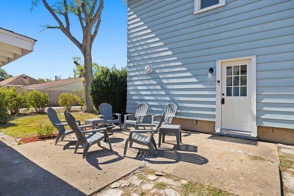 A patio with Adirondack chairs and a small table, a white door, and a tree in the background.