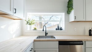 Cocina moderna con armarios blancos, pileta de acero inoxidable y lavavajillas. Se observa una ventana sobre la pileta con vista a árboles y una casa.