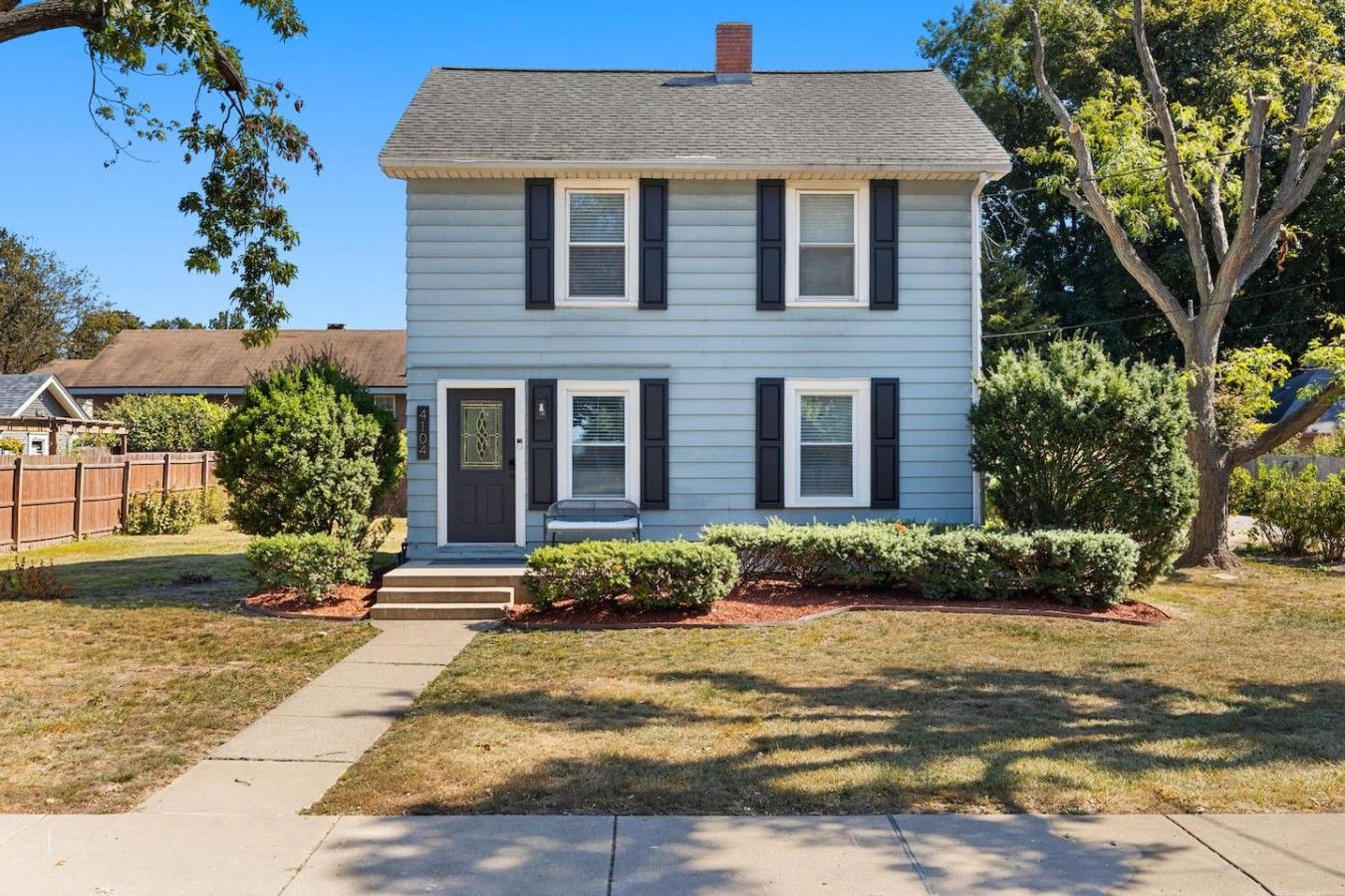 A two-story house with a front porch, a mailbox, and a small front yard with a tree and shrubs.
