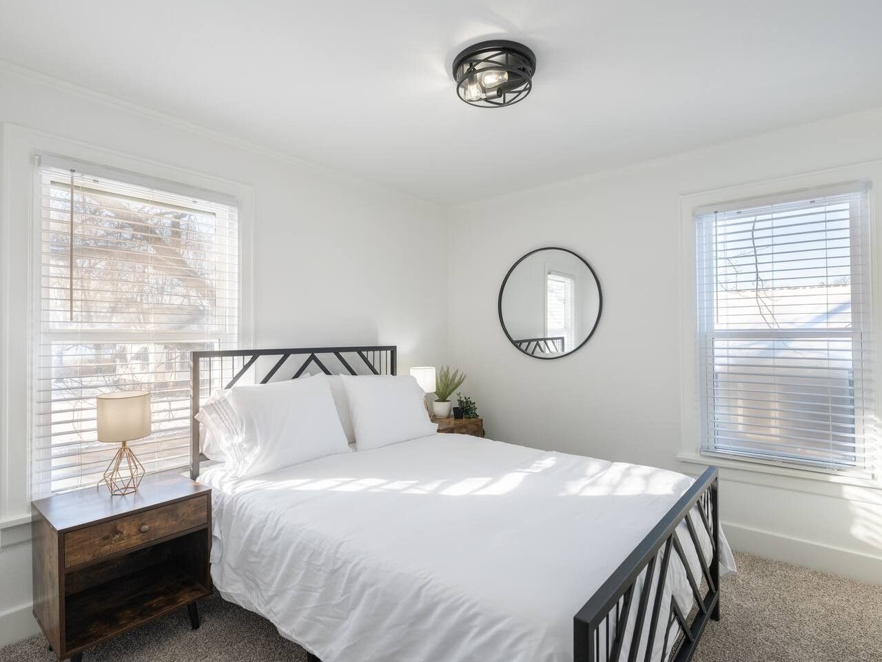 Une chambre à coucher comprenant un lit, une table de chevet, une lampe, un miroir et deux fenêtres équipées de stores.