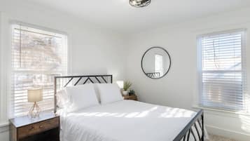 A bedroom with a bed, a nightstand, a lamp, a mirror, and two windows with blinds.