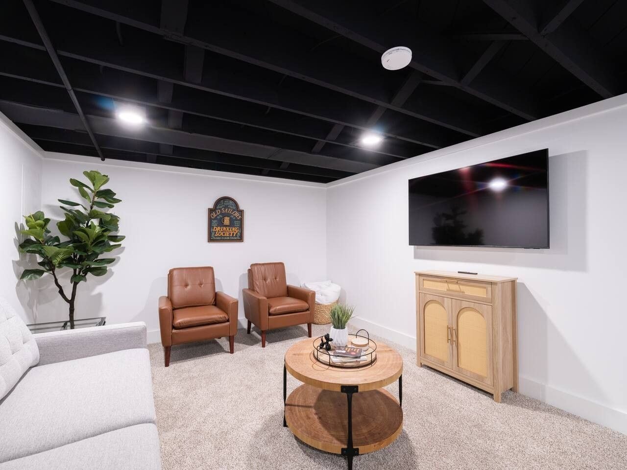 A modern living room with a flat-screen TV, two leather chairs, a round wooden coffee table, a white sofa, and a small potted plant.