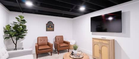 A modern living room with a flat-screen TV, two leather chairs, a round wooden coffee table, a white sofa, and a small potted plant.