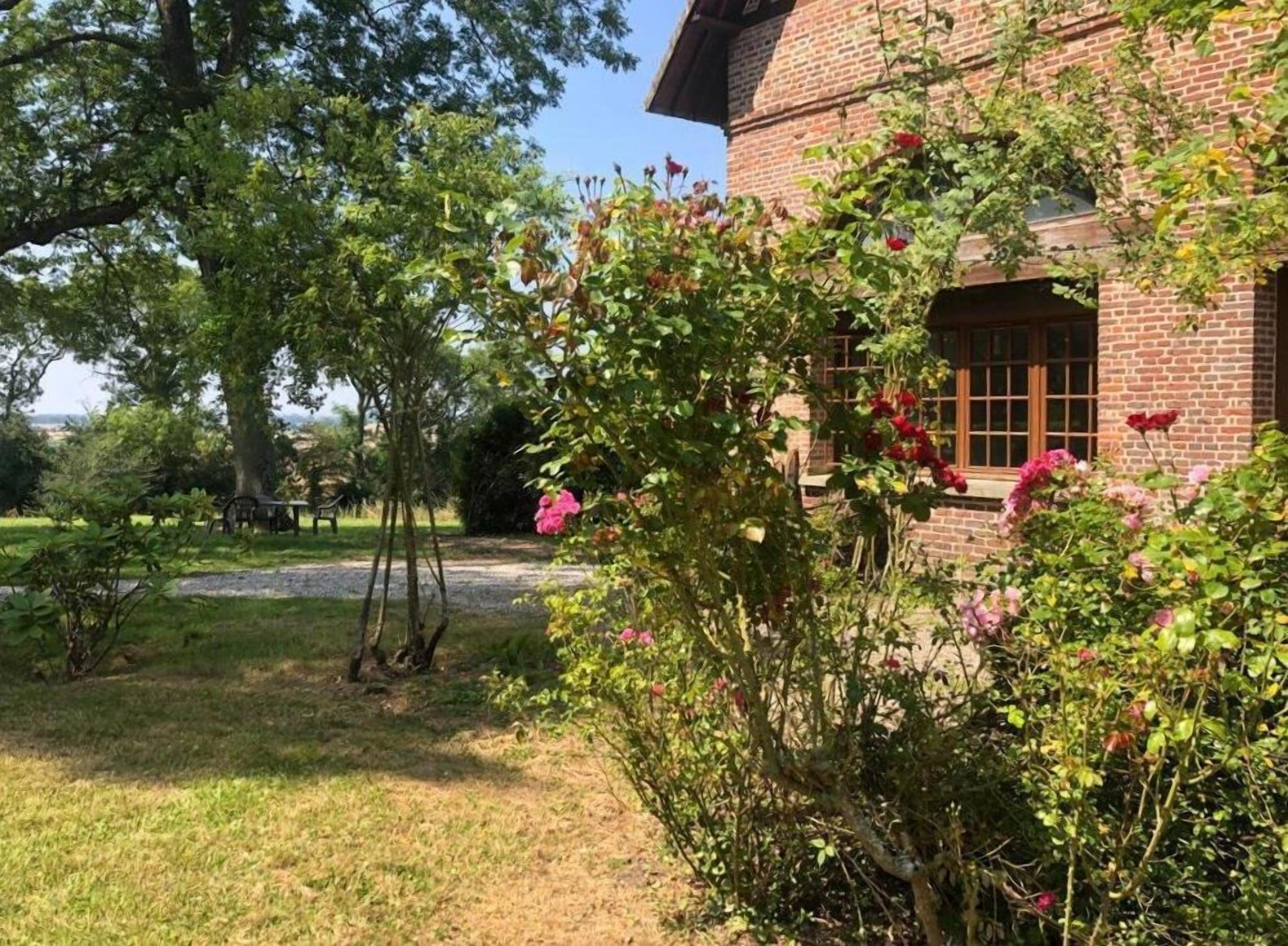 Un bâtiment en briques avec un jardin devant, comprenant un arbre et des arbustes en fleurs.