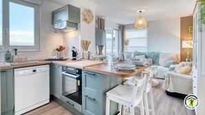 A modern kitchen with a dishwasher, oven, and a dining area with a table and chairs. - Gîtes de France® - Au Salon d'Arthur (CRIEL SUR MER)
