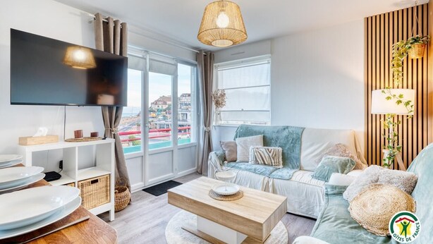 A modern living room with a flat-screen TV, a sofa, a coffee table, and a large window offering a city view. - Gîtes de France® - Au Salon d'Arthur (CRIEL SUR MER)