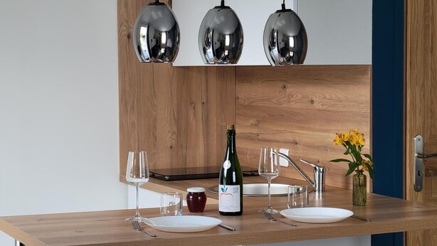 Une cuisine moderne avec des meubles de rangement en bois, un plan de travail en bois, trois lampes suspendues, une bouteille de vin et un vase rempli de fleurs.