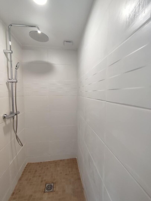 A small, white-walled shower room with a showerhead, handheld shower, and a floor drain.