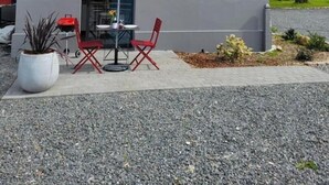 Un bâtiment moderne à l’extérieur gris, doté d’une petite fenêtre et d’un patio avec un parasol rouge, deux chaises et une table.