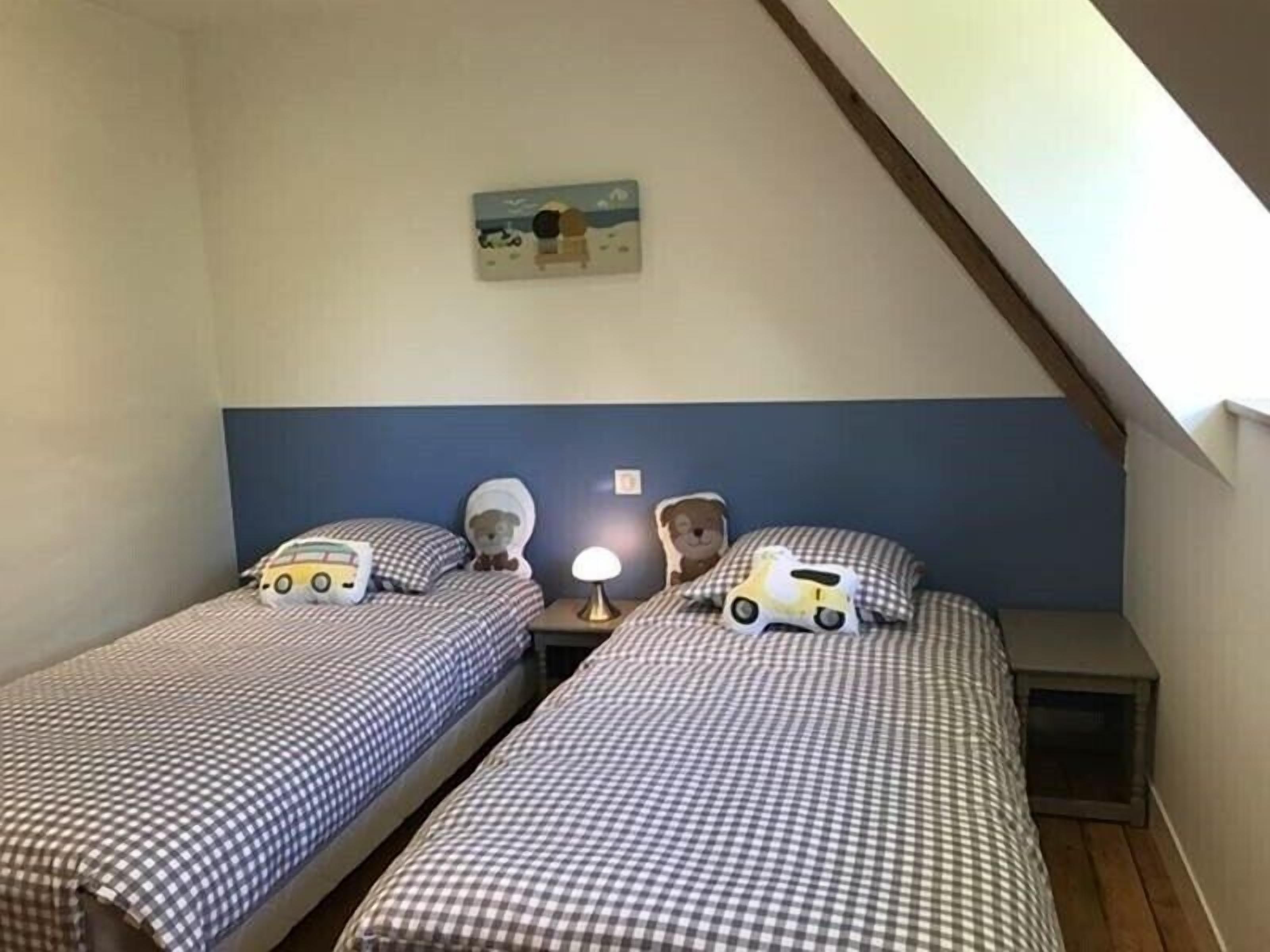 Une petite chambre accueillante avec deux lits, chacun agrémenté d’une couette à carreaux et d’un ours en peluche sur l’un des lits. Une table de chevet avec une lampe se trouve entre les deux lits, et une petite table avec une lampe est placée sur le côté droit. La chambre a un plafond en pente et un tableau est accroché au mur au-dessus des lits.