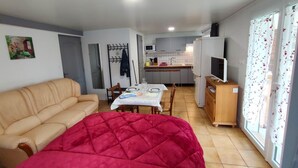 A compact living space with a sofa, a red quilted bed, a dining table with chairs, a kitchen area, and a television.