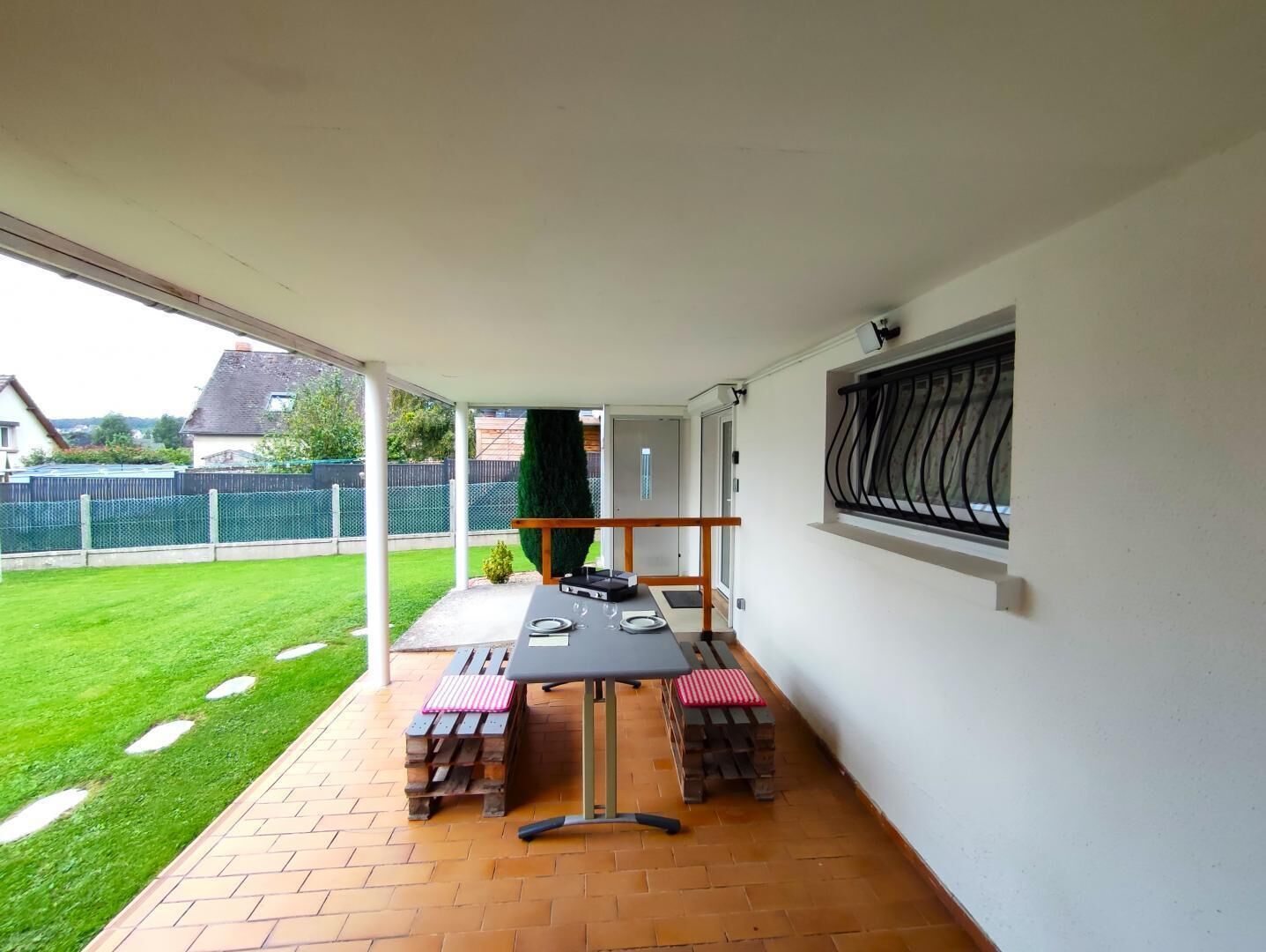 Uma varanda coberta com uma mesa posta para dois, uma janela com grade de metal e vista para um gramado verde e casas vizinhas.