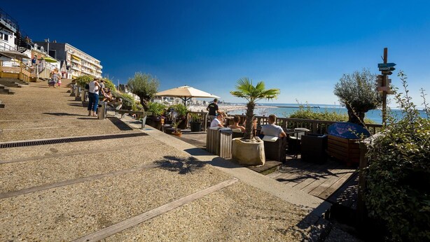 A seaside terrace with outdoor seating, a view of the beach, and a staircase leading down to the promenade.