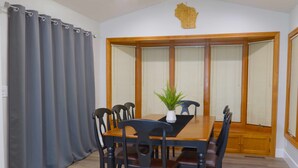 Une salle à manger avec une table en bois, des chaises noires, une plante en pot et une fenêtre avec des stores en bois.