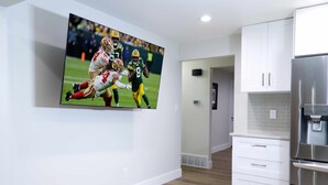Une cuisine moderne avec des meubles blancs, un réfrigérateur en acier inoxydable et un téléviseur à écran plat fixé au mur, diffusant un match de football.