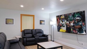 Un salon moderne avec un canapé en cuir noir, une table basse en bois et un grand téléviseur à écran plat diffusant un match de football.