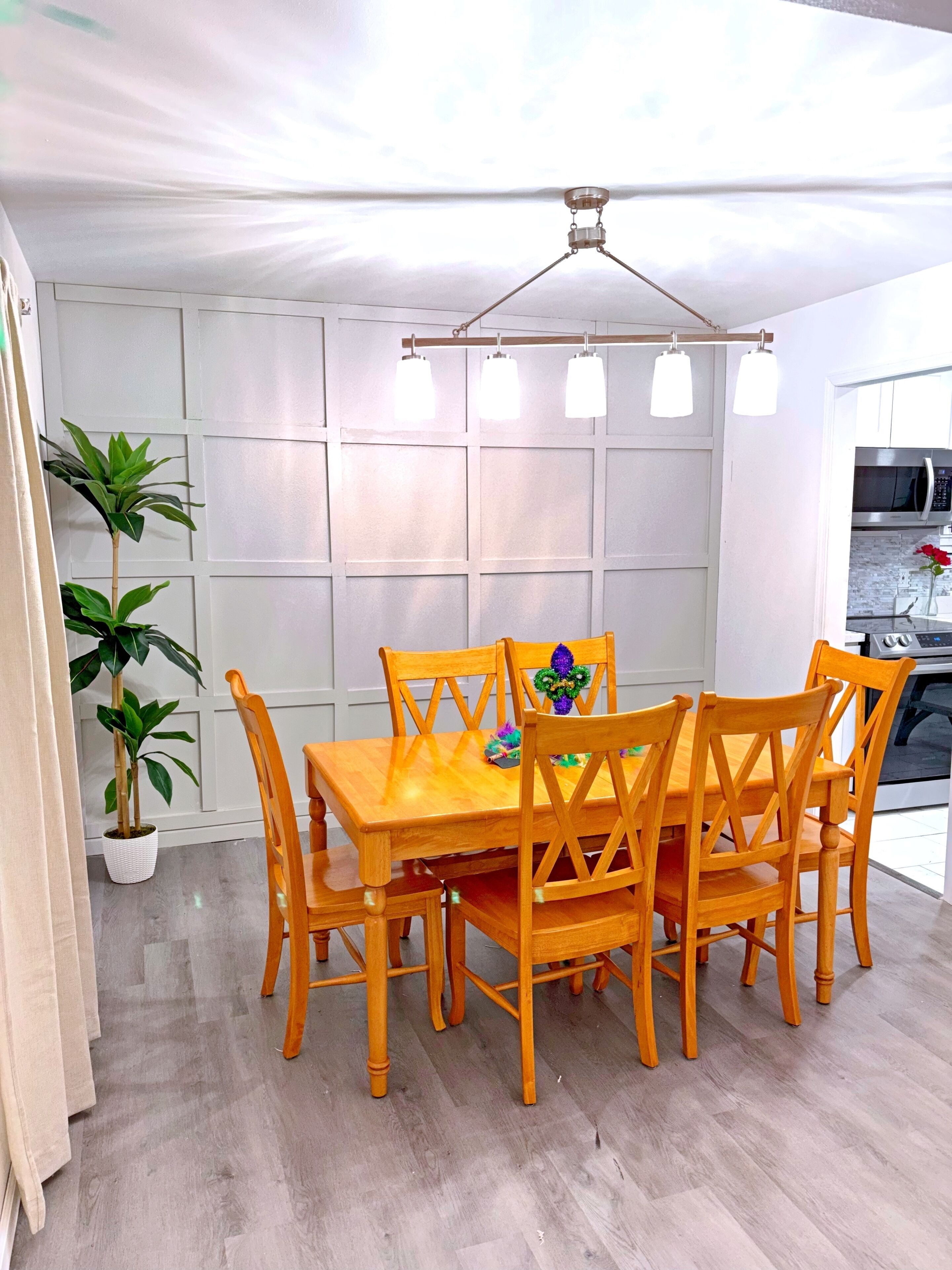 A dining area with a wooden table and chairs, a potted plant, and a modern kitchen in the background.