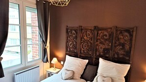 A bedroom with a large bed, a window with curtains, a bedside lamp, and a decorative headboard. - Gîtes de France® - Les Fées Mer (DIEPPE)