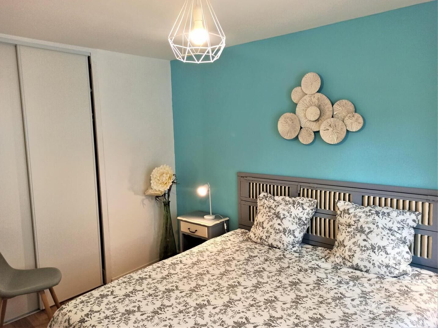 Une chambre à coucher comprenant un lit, une table de chevet, une lampe, une chaise et un élément décoratif mural.