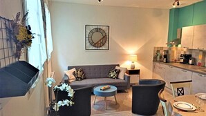 A compact living space with a sofa, two armchairs, a coffee table, and a dining table set for two. A lamp provides ambient lighting, and a clock is mounted on the wall. - Gîtes de France® - Les Fées Mer (DIEPPE)