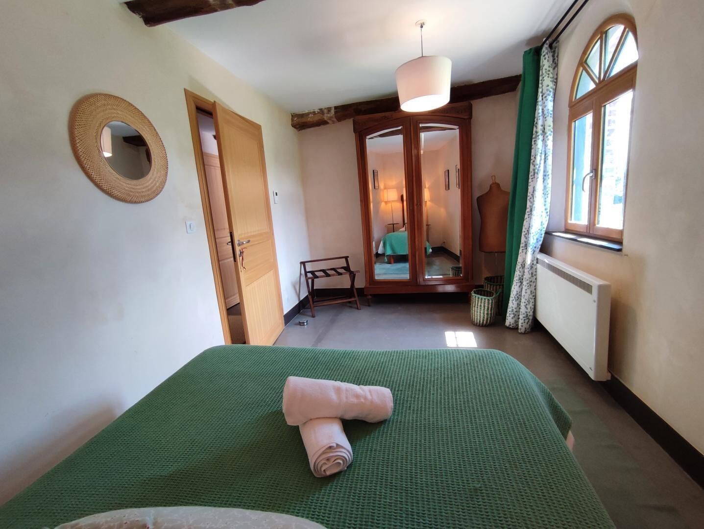 Een slaapkamer met een groen bedovertrek, een houten garderobe, een spiegel en een raam met gordijnen.