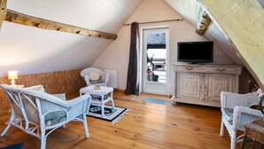 Una habitación cálida en el ático, con pisos de madera, muebles de mimbre blanco, un televisor y vistas por una puerta.