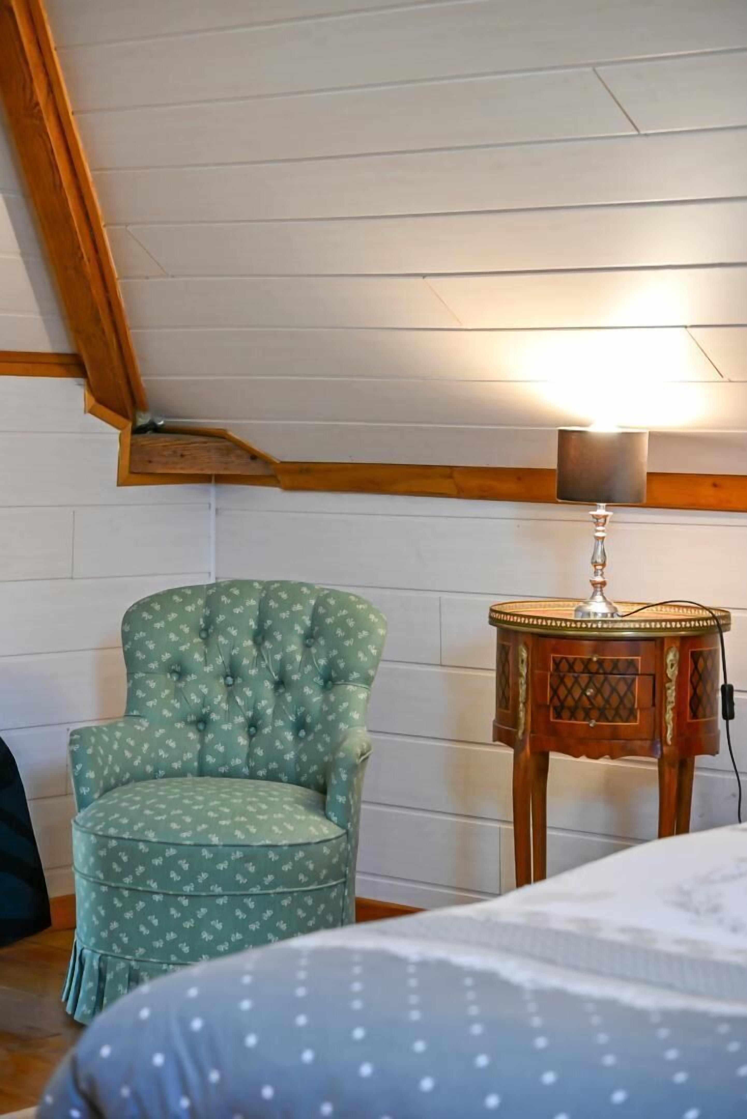 A room with a wooden ceiling, a green upholstered armchair, a small wooden side table with a lamp, and a bed with a white bedspread.