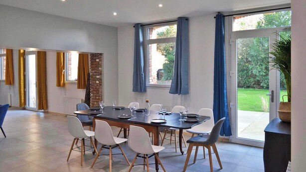 A dining area with a large table, chairs, and a view of a garden through the windows.