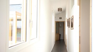 A bright, narrow hallway with large windows on one side, a wooden door at the end, and a ceiling light fixture.