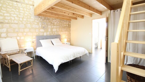 A bedroom with a stone wall, a bed with white bedding, a wicker chair, a nightstand, and a wooden ladder.