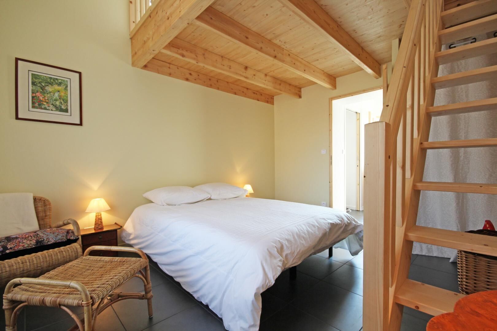 Un dormitorio con cama, mesita de noche, silla, escalera y un cuadro en la pared.