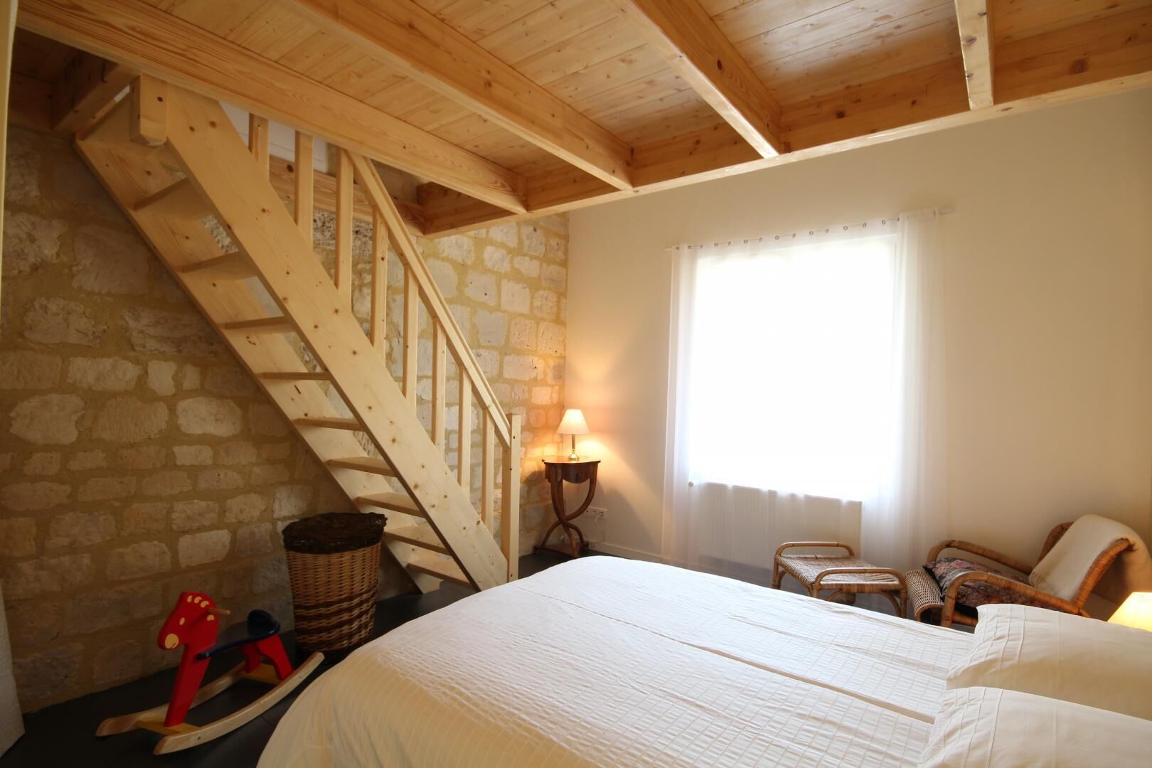 Ein Schlafzimmer mit einer Holztreppe, einem Bett, einem Fenster mit Vorhängen und einem kleinen Tisch mit einer Lampe.