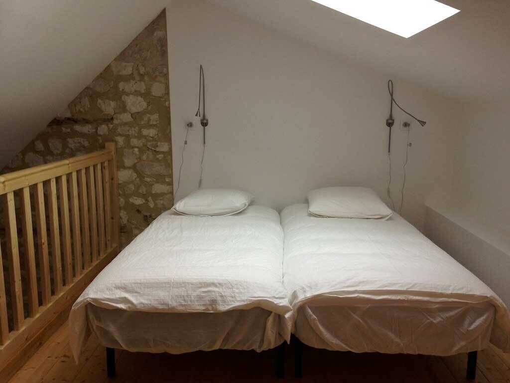 A small, single-room bedroom with a sloped ceiling and a stone wall. There are two beds with white bedding, a wooden railing, and a skylight.