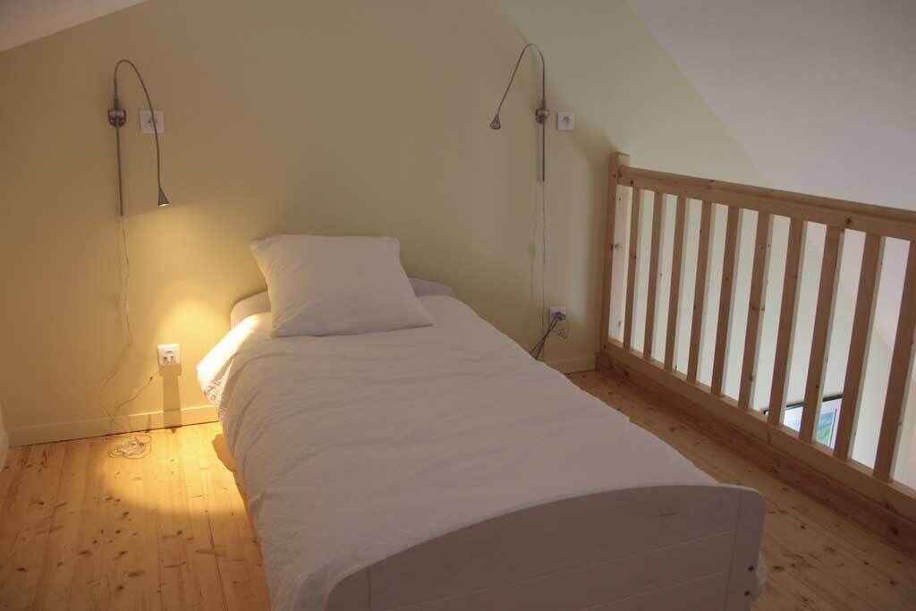 A small, single-bed room with a sloped ceiling and wooden flooring. There are two bedside lamps and a wooden railing along the window.