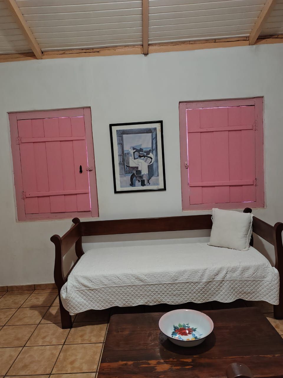 A room with a wooden sofa, a white quilted bedspread, a framed picture on the wall, and a bowl on a wooden table.