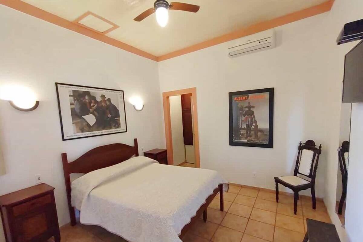 Un dormitorio con cama, mesitas de noche, una silla, ventilador de techo, luces fijadas a la pared y cuadros enmarcados.