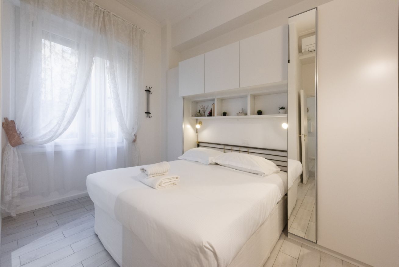 Ein Schlafzimmer mit Bett, Nachttisch, Spiegel und einem Fenster mit durchsichtigen Vorhängen.
