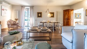 A living room with a sofa, ottoman, and a dining area with chairs and a table. - Villa Porri by MMega (Siena)