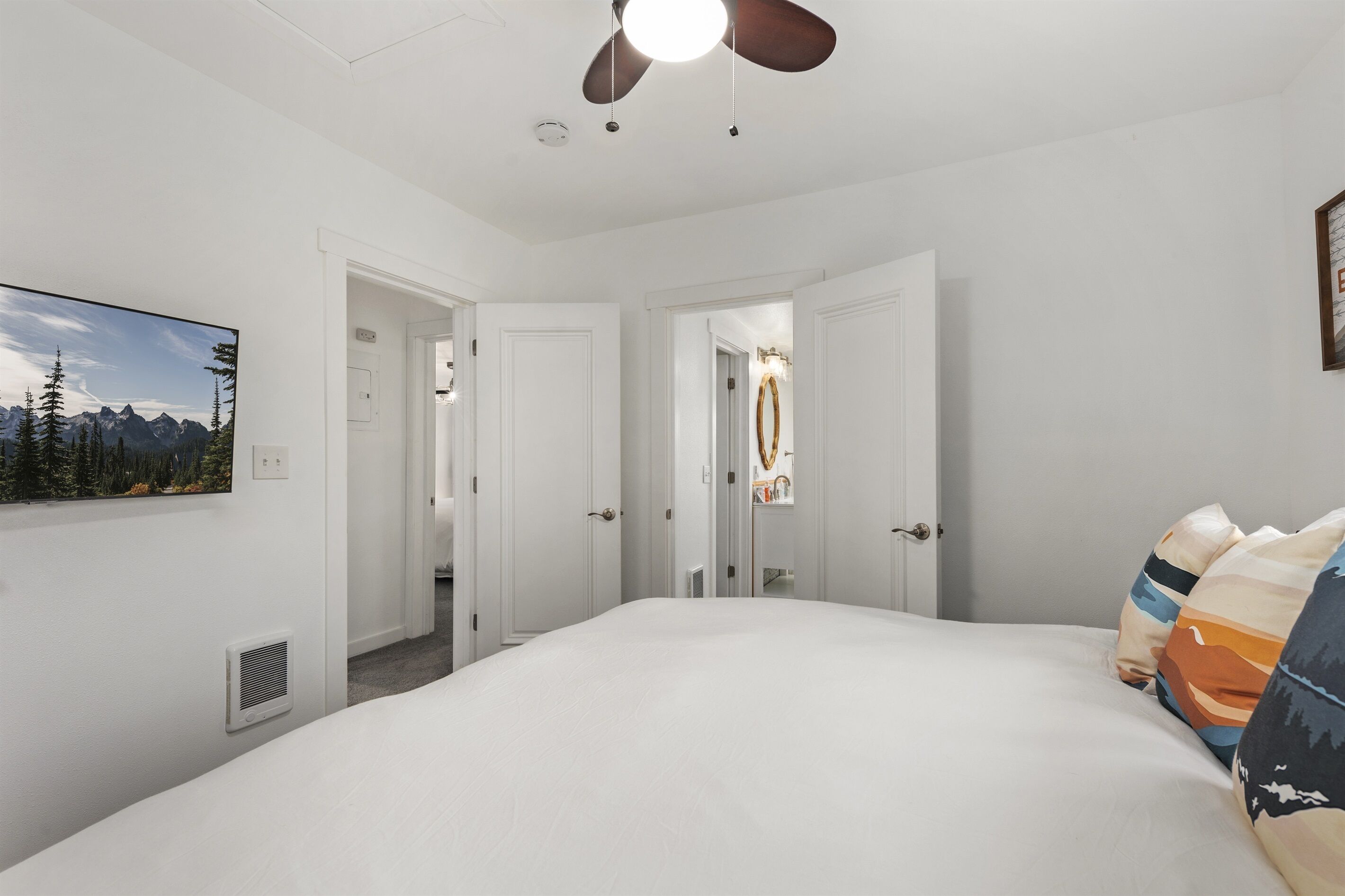 A hotel room with a large bed, a ceiling fan, a painting of mountains, and a door leading to another room.
