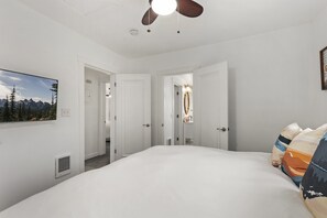 A hotel room with a large bed, a ceiling fan, a painting of mountains, and a door leading to another room.