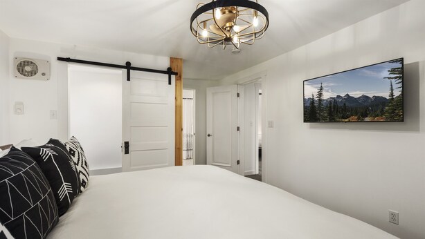 Un dormitorio con cama, televisor montado, lámpara de techo y puerta que da a otra habitación.