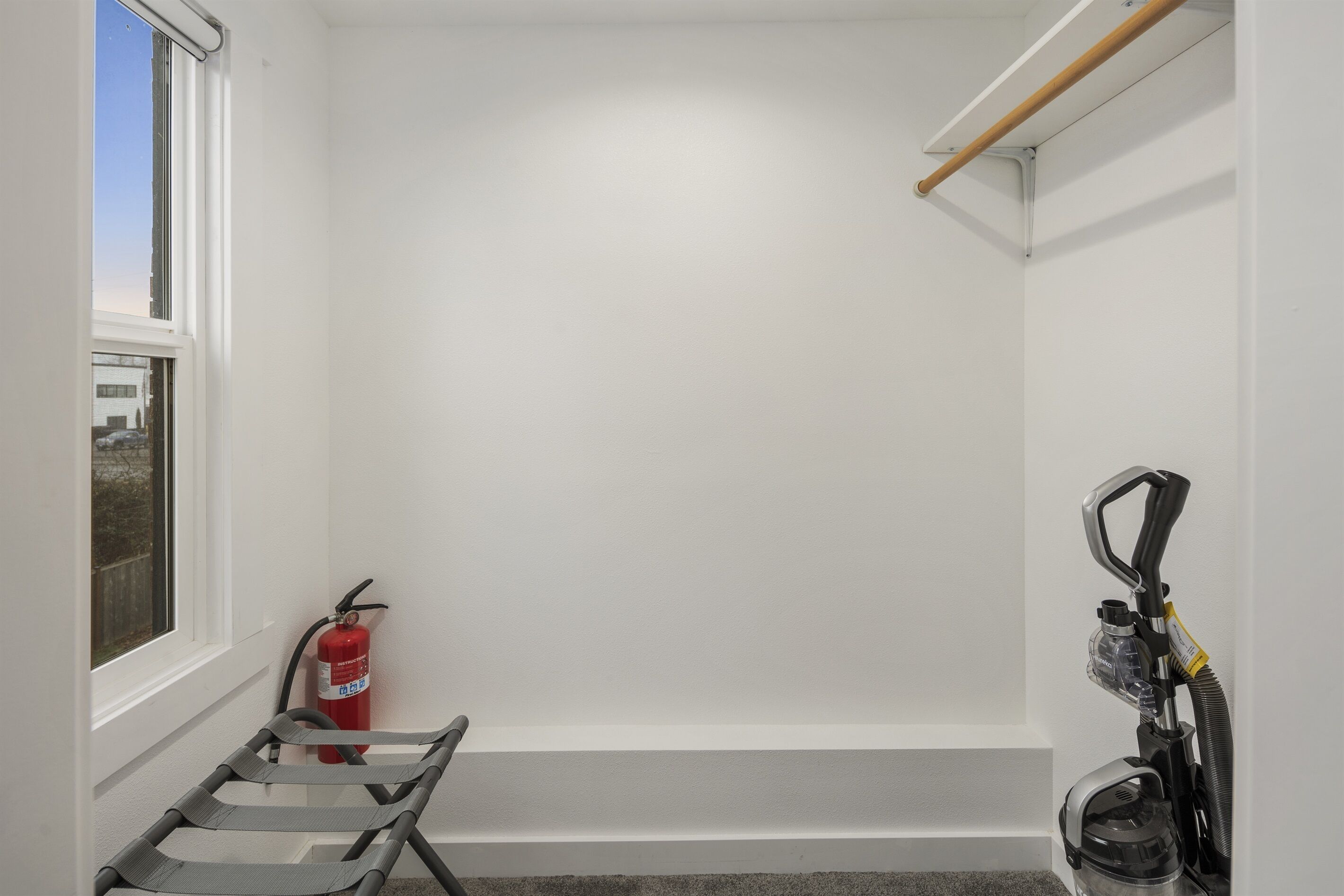 A corner of a room with a window, a fire extinguisher, a vacuum cleaner, and a wooden beam on the ceiling.