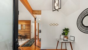 Un interior moderno con piso de madera, una pared blanca con patrones geométricos, un espejo redondo y una pequeña mesa con una planta y certificados enmarcados.