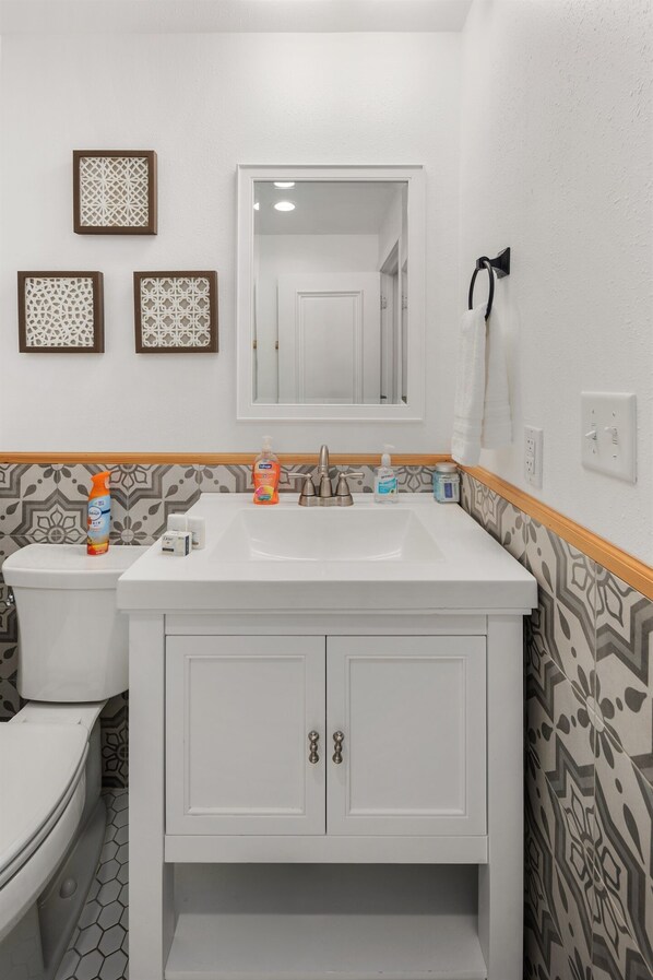 Un baño con lavamanos, espejo y retrete blancos. Se observan paneles decorativos en la pared y un piso estampado.