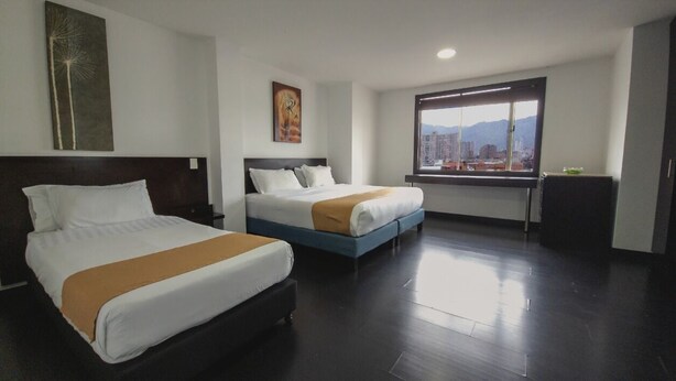 A hotel room with two beds, a large window with a city view, and a painting on the wall. - Hotel City Center Corferias (Bogotá)